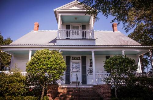 A charming two-story white house with a porch, surrounded by greenery and a clear blue sky.
