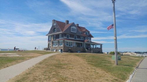 A large, historic stone house with a red roof, set against a blue sky, near a grassy area and a flagpole.