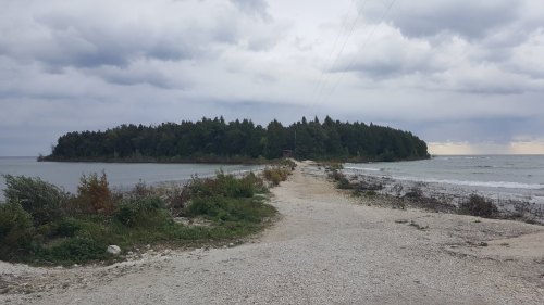 A gravel path leads to a small island surrounded by water, with cloudy skies and trees in the background.