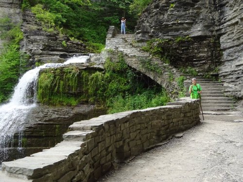 A scenic waterfall with stone steps and a hiker in green, surrounded by lush greenery and rocky cliffs.