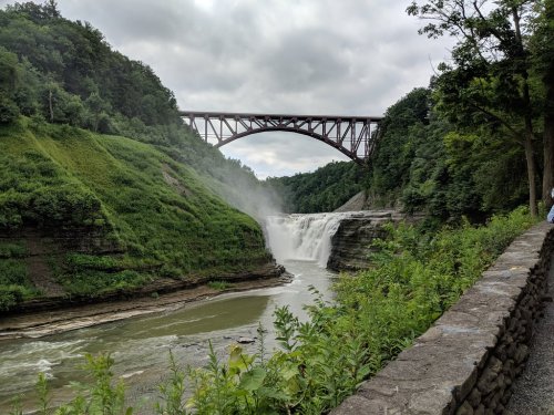 A scenic view of a waterfall beneath a bridge, surrounded by lush greenery and a cloudy sky.