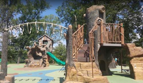 A nature-themed playground with wooden structures, slides, and climbing features surrounded by trees.