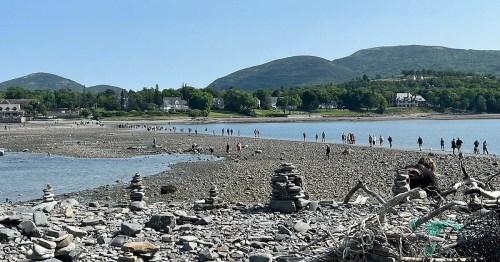 A rocky beach with people walking along the shore and stacked stones in the foreground, surrounded by green hills.