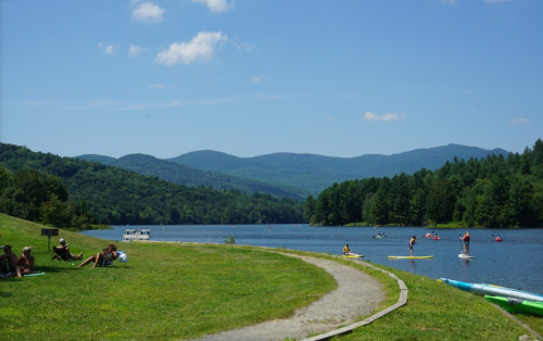 A serene lake scene with people paddleboarding, lush green hills, and a clear blue sky.