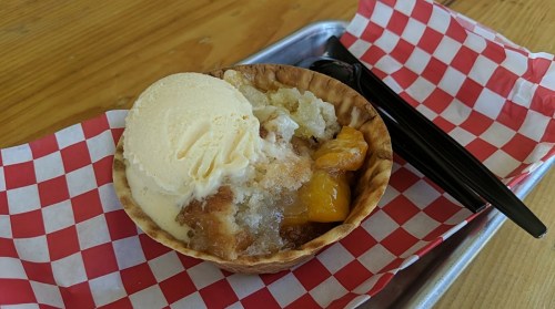 A dessert bowl with peach cobbler topped with vanilla ice cream, served on a red and white checkered paper.