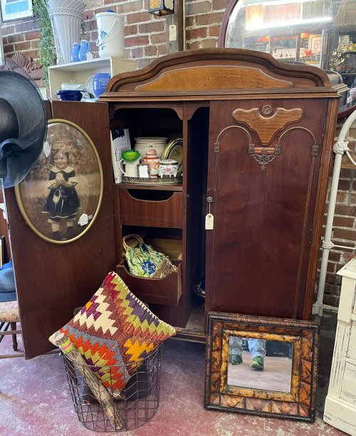 A vintage wooden cabinet with open doors, displaying various items, a framed photo, and a colorful patterned pillow.