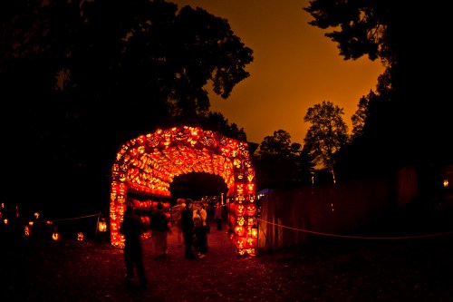 A glowing archway made of carved pumpkins, with people walking underneath against an orange sky.