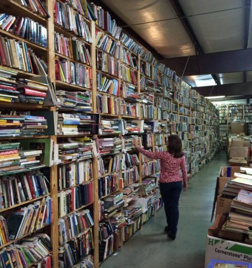 A person walks through a narrow aisle between tall shelves filled with books in a large, organized bookstore.