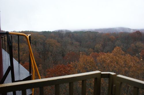 View from a balcony overlooking a misty forest with autumn foliage and distant hills under cloudy skies.