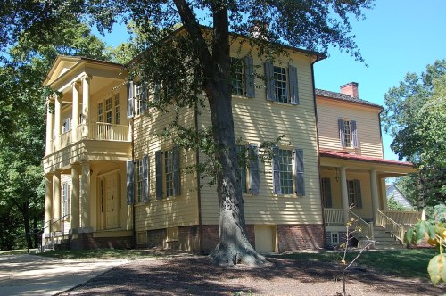 A historic yellow house with a porch, surrounded by trees and greenery on a sunny day.