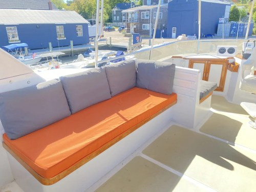 A boat interior featuring a cushioned bench with gray and orange cushions, overlooking a marina with boats.