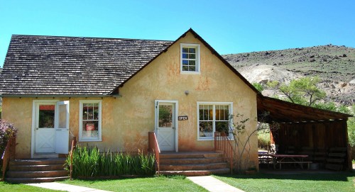 A charming, rustic house with a sloped roof, surrounded by greenery and mountains under a clear blue sky.