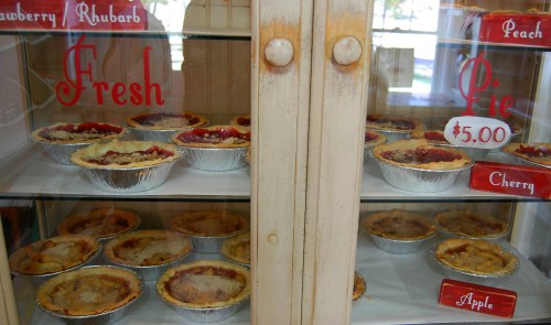 A display case filled with various fresh pies, labeled with flavors like strawberry rhubarb, peach, cherry, and apple.