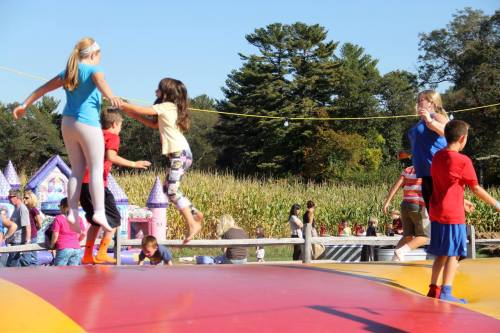 Children jumping on a colorful inflatable bounce pad, with a festive background of trees and a cornfield.