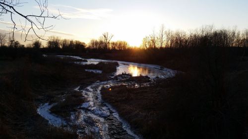 A serene sunset over a winding river, surrounded by trees and grassy banks, reflecting warm golden hues.