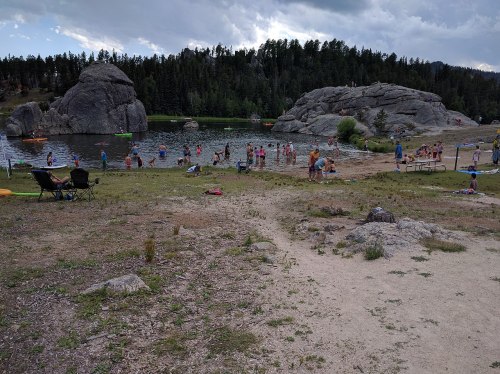 A scenic lake with people swimming and relaxing on the shore, surrounded by large rocks and trees under a cloudy sky.