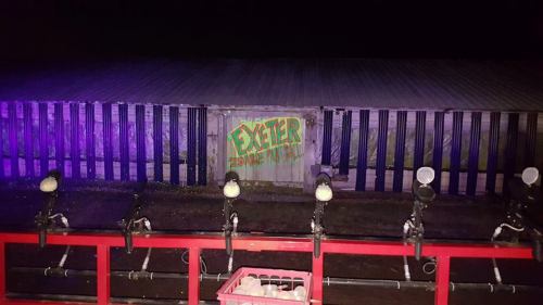A dimly lit scene featuring a wooden structure with "EXETER" spray-painted on it, viewed from a red railing with lights.