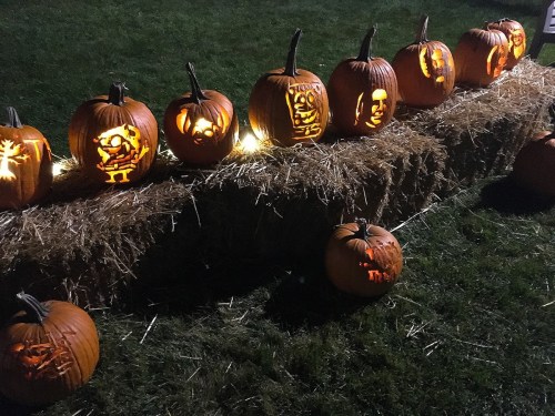 A row of carved pumpkins with various designs, illuminated and placed on a hay bale in a grassy area.