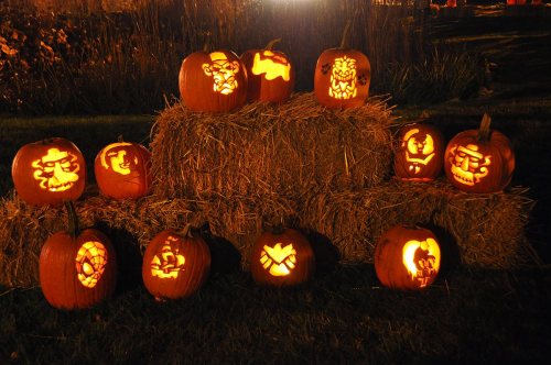 A display of carved pumpkins with various designs, illuminated and arranged on hay bales at night.