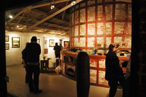 A cozy art gallery with brick walls, featuring visitors admiring paintings and a unique circular display area.