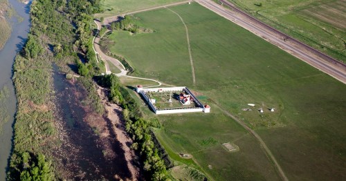 Aerial view of a historic site surrounded by green fields and a river, with pathways and structures visible.