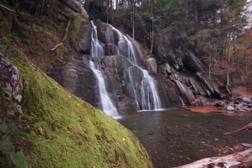 A serene waterfall cascades over moss-covered rocks, surrounded by lush green trees and a tranquil pool below.