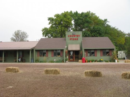 A green country store with a red roof, surrounded by hay bales and trees, set in a rural landscape.