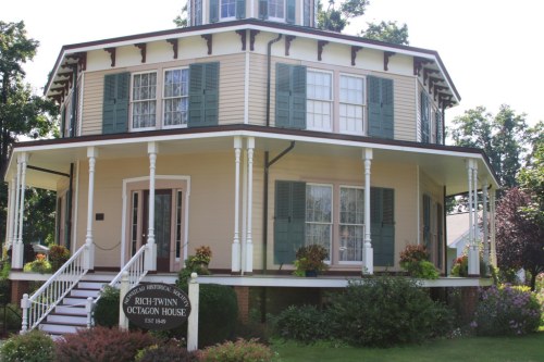 A historic octagon house with green shutters, white railings, and colorful flower beds, surrounded by lush greenery.