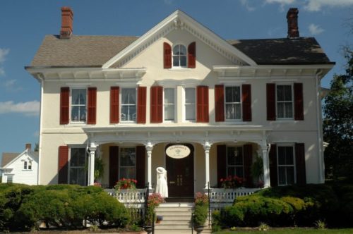 A large, elegant white house with red shutters and a porch, surrounded by greenery and flowers under a blue sky.