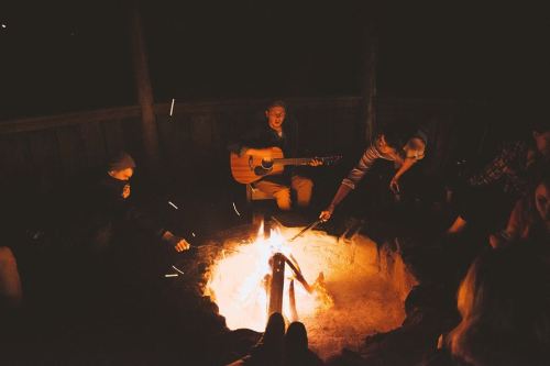 A group of people gathered around a campfire at night, with one person playing guitar and others enjoying the warmth.