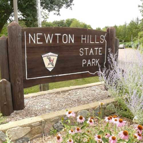 Sign for Newton Hills State Park, featuring a logo and surrounded by greenery and flowers.