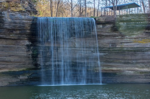 A serene waterfall cascading over a rocky cliff into a calm pool, surrounded by trees and a shelter above.