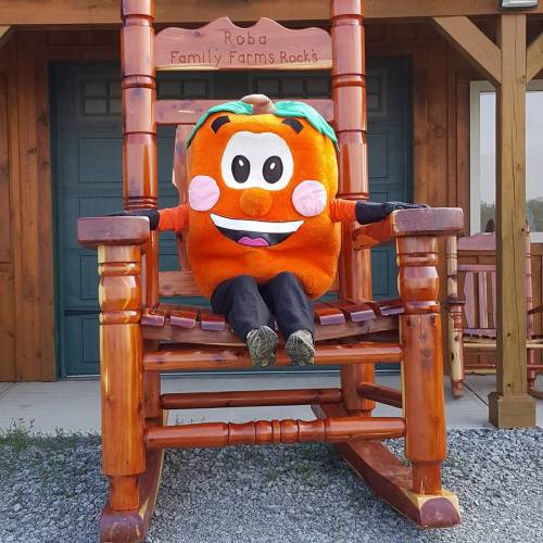 A large orange mascot sits in a wooden rocking chair at a farm, smiling and wearing blue pants.