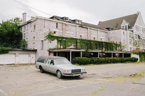 An old hearse parked in front of an abandoned, overgrown building with peeling paint and a cloudy sky.
