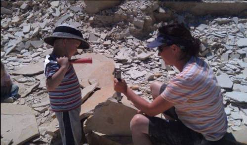 A child with a hammer watches as an adult chisels a rock in a rocky outdoor setting.