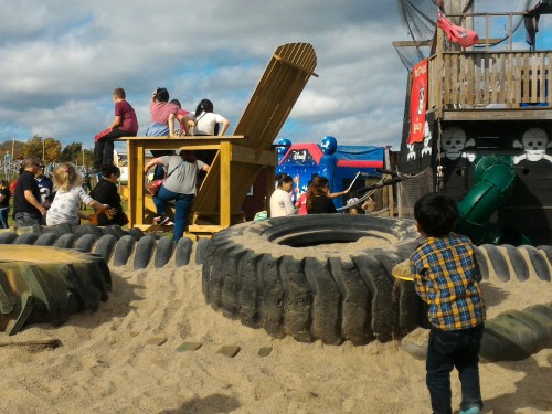 Children play in a sandy playground with large tires, a wooden structure, and pirate-themed decorations in the background.