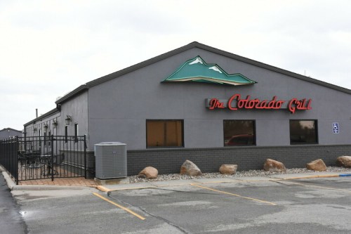 Exterior of The Colorado Grill, featuring a mountain logo, large windows, and a fenced patio area.