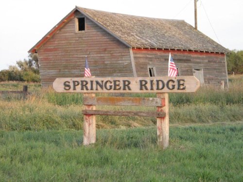 A wooden sign reading "Springer Ridge" stands in front of a weathered barn, with American flags on either side.