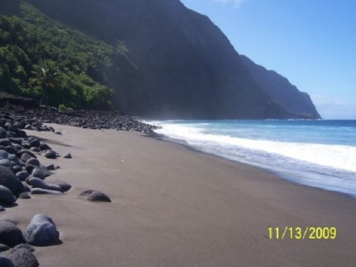 A serene beach with black sand, smooth stones, and lush green mountains under a clear blue sky.