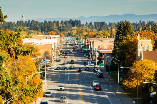 A busy street lined with trees in autumn colors, with cars and buildings under a clear blue sky and distant mountains.