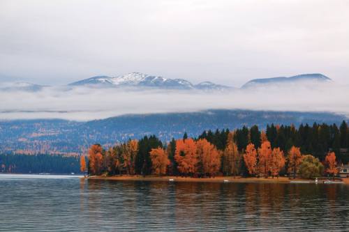 A serene lake surrounded by autumn trees with vibrant orange leaves, mountains in the background, and misty clouds.