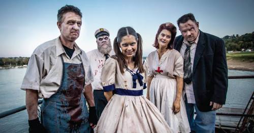 A group of five people in horror-themed costumes, featuring blood and eerie expressions, by a lakeside.