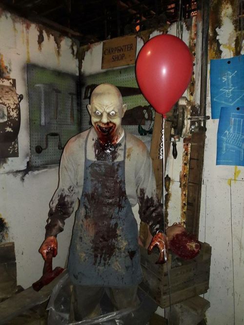 A creepy, bloodied mannequin in an apron holds a red balloon, set in a dimly lit workshop with eerie decor.