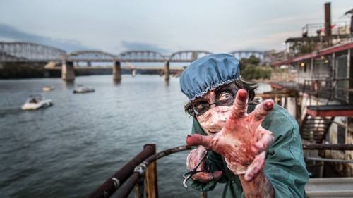 A creepy figure in a surgical outfit and mask reaches out, with a river and bridge in the background.