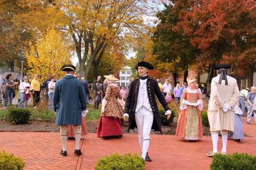 A group of people in historical costumes walking in a park with autumn foliage and a crowd in the background.