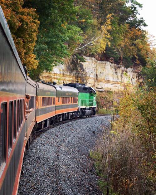A train curves around a rocky cliff, surrounded by colorful autumn foliage.