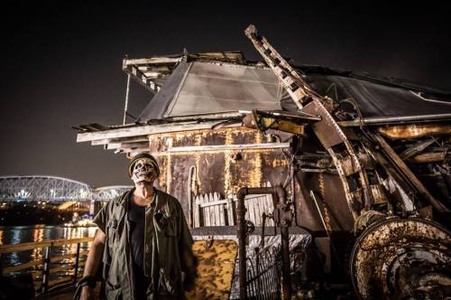 A person in a skull mask stands near a rusty, abandoned structure at night, with a bridge visible in the background.