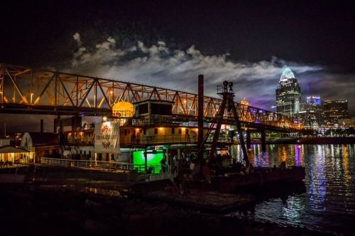 Nighttime view of a city skyline with a bridge over water, featuring illuminated buildings and a lively waterfront scene.