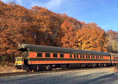A vintage train car in green and orange parked beside vibrant autumn foliage.