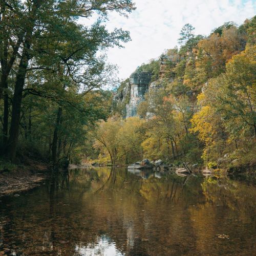 A serene river flows through a forested landscape, with colorful autumn trees and rocky cliffs in the background.